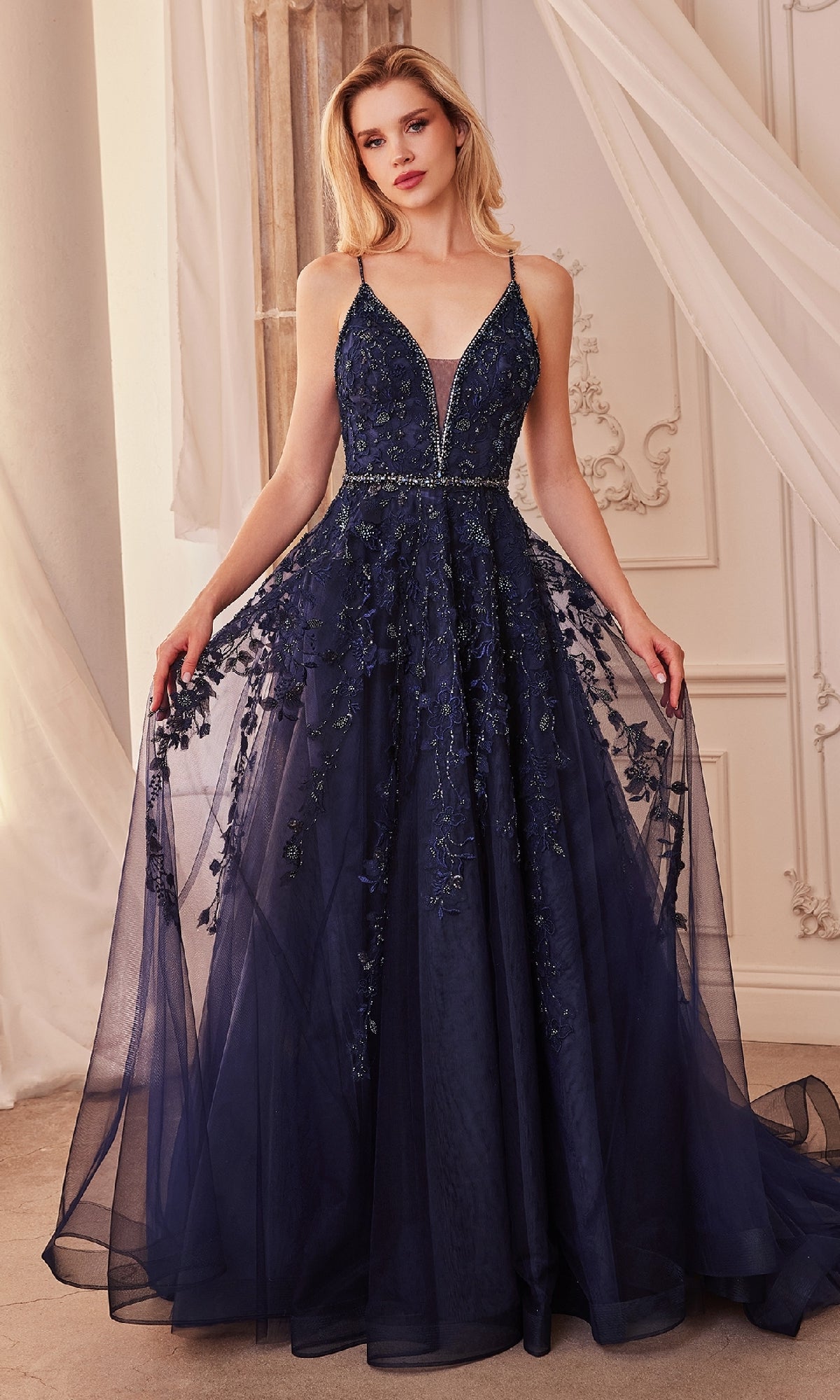 A woman stands indoors wearing the Long Formal Dress A1251 by Andrea and Leo—a navy blue ball gown featuring a floor-length sheer overlay, floral embroidery, thin straps, and an elegant v-neckline.